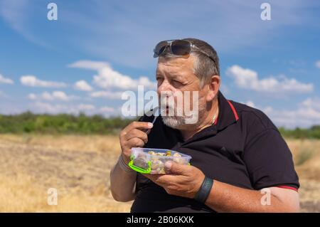 Porträt des kaukasischen Bärtigen Senior beim Essen von gedämpftem Gemüse im Freien Stockfoto