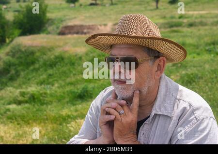 Außenporträt des fröhlichen ukrainischen Landsmannes in Strohhut und Sonnenbrille gegen die hüglige Quellweide Stockfoto