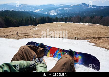 Blick auf Snowboarder und buntes Snowboard im Schnee, Blick auf die Winterlandschaft. Bergrichtung unter hohen Fichten, verschneite Gipfel im Hintergrund. Sport- und Pov-Konzept. Stockfoto