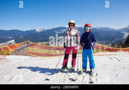 Zwei Teenager, Junge und Mädchen in warmer Kleidung und Brille auf Skiern im Tiefschnee vor dem Hintergrund des Skigebietes, des leuchtend blauen Himmels und der Winterberge. Sport-, Freizeit- und Outdoor-Aktivitäten Konzept. Stockfoto