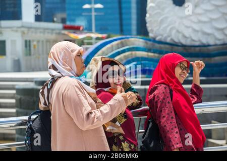 Drei indonesische Touristen tragen Hijab und posieren mit einem großen, glücklichen Lächeln für ein Gruppenfoto vor dem Merlion Statue Park. Singapur. Stockfoto