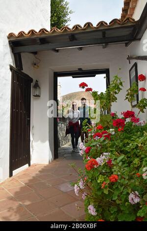 Junges, attraktives lächelndes Paar, das durch die offene Tür von weiß getünchten Gebäuden in Betancuria, Fuerteventura, geht. Blumen im Vordergrund. Stockfoto