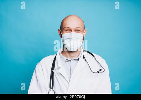 Attraktiver freundlicher kahlbärtiger lächelnder Arzt in medizinischer Maske mit Blick auf die auf blauem Hintergrund isolierte Kamera Stockfoto
