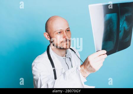 Attraktiver freundlicher kahler lächelnder Arzt, der die auf blauem Hintergrund isolierte Röntgenaufnahme betrachtet Stockfoto