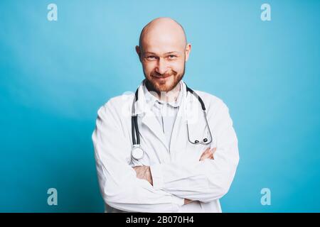 Attraktiver freundlicher kahler lächelnder Arzt, der die Kamera isoliert auf dem blauen Hintergrund betrachtet Stockfoto