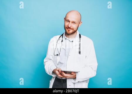 Attraktiver freundlicher, kahler lächelnder Arzt mit Tablette, die auf dem blauen Hintergrund isoliert auf die Kamera blickt Stockfoto