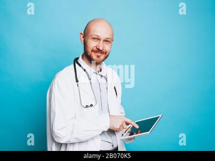 Attraktiver freundlicher, kahler lächelnder Arzt mit Tablette, die auf dem blauen Hintergrund isoliert auf die Kamera blickt Stockfoto