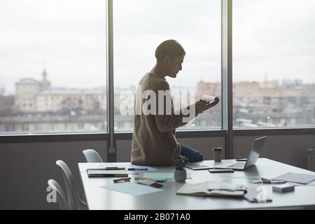 Junger Geschäftsmann, der auf dem Tisch sitzt und den Bericht vor dem Treffen im Büro prüft Stockfoto