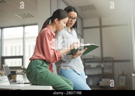 Zwei junge Geschäftsfrauen in legerer Kleidung mit Notizblock und Blick auf ihre Pläne für eine Arbeitswoche im Büro Stockfoto
