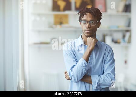 Penibler afrikanischer Geschäftsmann in Brillen, die im Büro stehen und wegschauen, denkt er über etwas nach Stockfoto