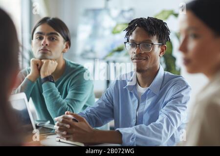 Junger afrikanischer Geschäftsmann in Brillen, die am Tisch sitzen und seinen Kollegen bei der Sitzung im Büro zuhören Stockfoto