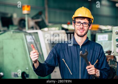 Junger Arbeiter mit einem Reparaturingenieur repariert die Maschine glücklich lächelnd in der Werkshäube, um ein gutes Arbeitssignal zu erhalten Stockfoto