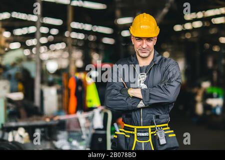 Fröhlicher Arbeiter, Portrait schöne Arbeitsarme gefaltet mit Sicherheitsanzug Werkzeuggürtel und Funkdienst Mann in der Fabrik. Stockfoto