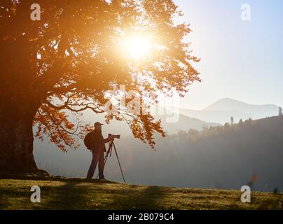 Männlicher Touristenfotograf mit Rucksack, Stativ und professioneller Kamera unter großer Eiche mit goldenem Laub auf holziger Nebelberge Landschaft und heller Sonne im blauen Himmel Hintergrund. Stockfoto