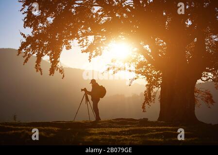 Profil des Reisenden Wanderer Mann mit Rucksack auf Kamerastativ beleuchtet von der hellen untergehenden Sonne, die auf grassem Tal steht, auf dem Hintergrund des schönen Bergpanoramas in der Dämmerung unter großem Baum. Stockfoto