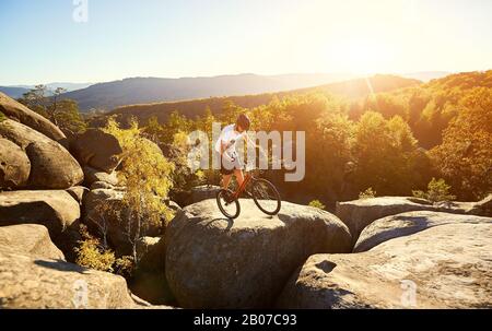 Sportler Radfahrer balanciert auf dem Hinterrad auf dem Trial Bike, Sportlerfahrer macht bei Sonnenuntergang akrobatische Stunt am Rande eines großen Felsboulders auf der Spitze des Berges. Konzept des aktiven Lifestyles des Extremsports Stockfoto