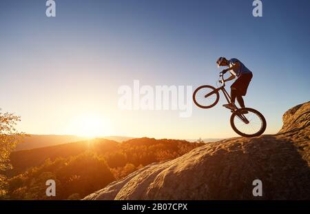 Silhouette des Radfahrers, der auf dem Hinterrad auf dem Trial-Fahrrad balanciert. Mutiger Sportbiker, der bei Sonnenuntergang auf dem felsigen Berg akrobatischen Trick macht. Konzept des aktiven Lifestyles des Extremsports Stockfoto