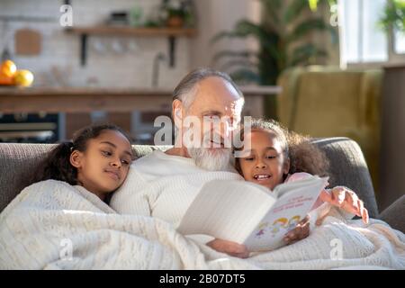 Grauhaariger Großdad liest Schlafensgeschichten seinen Enkeltöchtern vor Stockfoto
