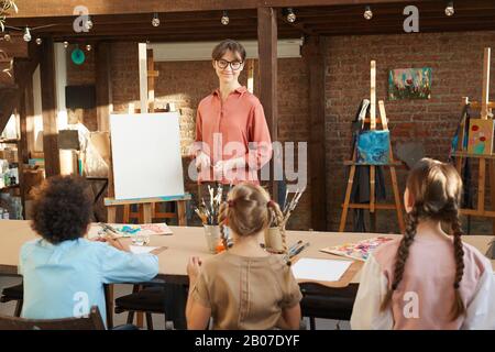 Junge Lehrerin, die Kindern beigebracht hat, zu malen, während sie während des Kunstunterrichts im Kunststudio am Tisch sitzen Stockfoto