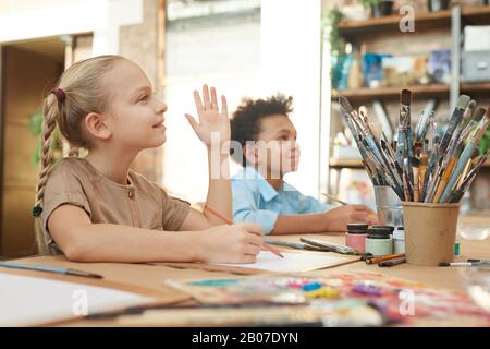 Kleine Mädchen, die ihre Hand heben, während sie am Tisch sitzen und während des Kunstunterrichts mit einem Jungen im Hintergrund malen Stockfoto
