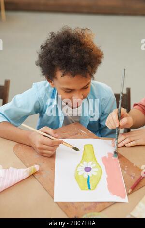 Afrikanischer kleiner Junge, der während des Kunstunterrichts am Tisch sitzt und ein Bild mit Farben malt Stockfoto