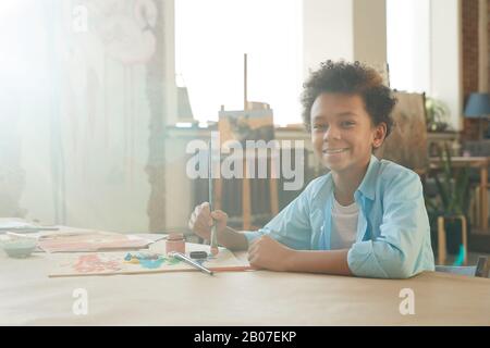 Porträt des afrikanischen jungen Jungen, der beim Sitzen am Tisch mit der Kamera lächelt und ein Bild mit Farben und Pinsel zeichnet Stockfoto