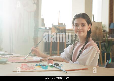 Porträt des kleinen Mädchens, das die Kamera betrachtet, während er ein Bild am Tisch im Kunststudio malt Stockfoto