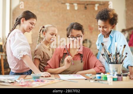 Junge Lehrerin in Brillen, die am Tisch sitzen und zeigen, wie sie ihren Schülern im Kunststudio ein Bild zeichnet Stockfoto