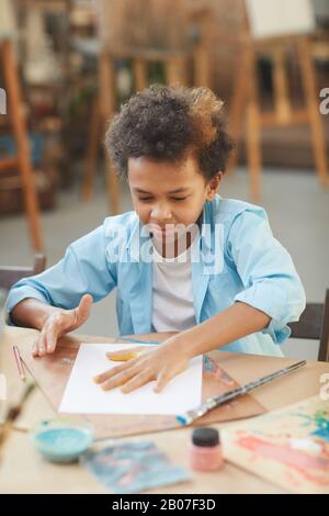 Afrikanischer kleiner Junge, der am Tisch sitzt und im Kunststudio eine Handarbeit mit Farben macht Stockfoto