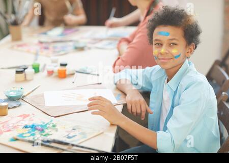 Porträt eines afrikanischen Jungen, der mit Papier und Farben bei Kunstunterricht am Tisch sitzt und die Kamera betrachtet Stockfoto