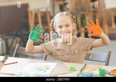Porträt eines glücklichen Mädchens, das mit der Kamera lächelt und ihre Hände in Farben zeigt, während sie im Kunststudio am Tisch sitzt Stockfoto