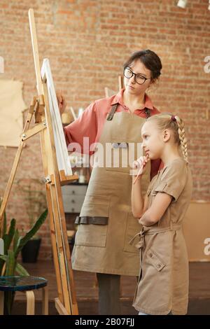 Junge Lehrerin, die mit einem kleinen Mädchen in der Nähe der Staffelei steht und ihr zeigt, wie sie während des Kunstunterrichts im Studio ein Bild malt Stockfoto