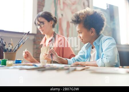 Junge Frau, die zusammen mit dem afrikanischen Jungen am Tisch sitzt und im Kunststudio mit Aquarellen malt Stockfoto