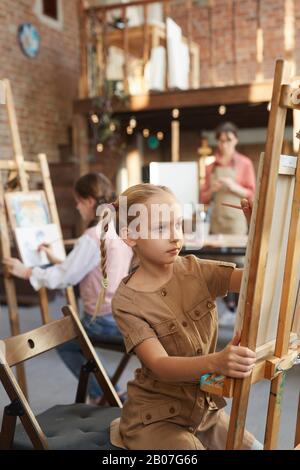 Kleines Mädchen, das während des Kunstunterrichts im Kunststudio vor der Staffelei sitzt und mit anderen Studenten malt Stockfoto