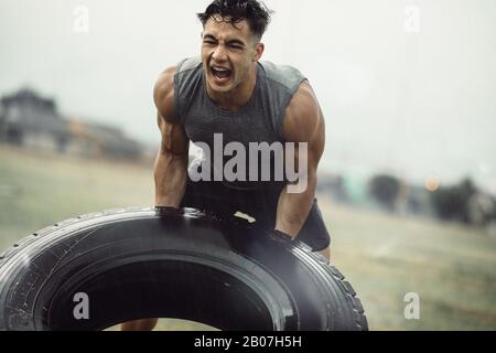 Harter junger Athlet, der im Regen eine Übung zum Spiegeln des Reifens macht. Muskulöser Mann macht Cross-Training im Freien auf dem Feld. Stockfoto