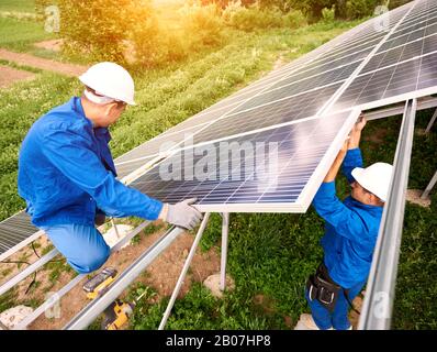 Installation eines Standalone-Solarfoto-Voltaic-Panel-Systems. Zwei Techniker in Harthüten montieren großes glänzendes Solarmodul auf Plattform auf grünem Hintergrund im Sommer. Alternatives Energiekonzept. Stockfoto