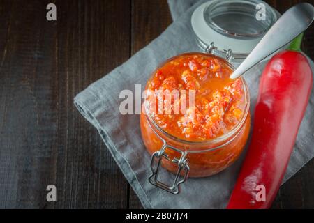 Traditionelle Adjika heiße Chili Pfeffer Soße Paste Harissa in Glas Glas auf dunkel. Tunesien-Küche, georgische und arabische Küche. Horizontale Ausrichtung. Platz für Stockfoto