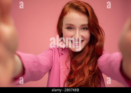 Nahaufnahme des Porträts der schönen jungen Rotkopffrau, die selfie mit zwei Händen macht. Lustige lächelnde Mädchen mit pinkfarbener Jacke, die ein Geschenk auf dem Stift isoliert Stockfoto