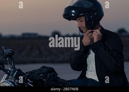 Lateinischer Mann, der seinen Helm aufsetzt, um sein Motorrad bei Sonnenuntergang zu fahren. Stockfoto