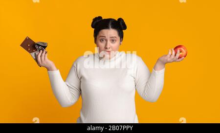 Verwirrte Übergewichtige Frau, Die Chocolate Bar Und Apple In Händen Hält Stockfoto
