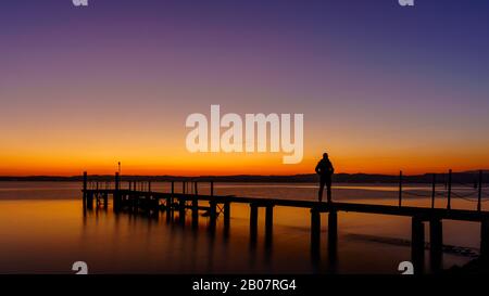 Eine Mannsilhouette, die auf einem Holzsteg einsam am Meer mit wunderschönem Sonnenuntergang steht. Lsunset seekape an einem Holzsteg. Stockfoto