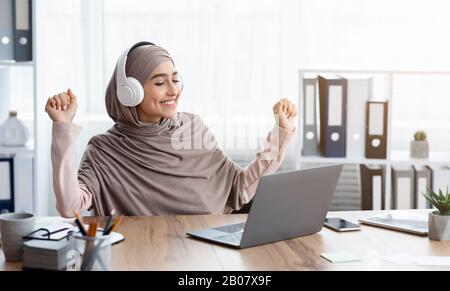 Freudige arabische Frau in Hijab, die Musik in Kopfhörern am Arbeitsplatz hört Stockfoto