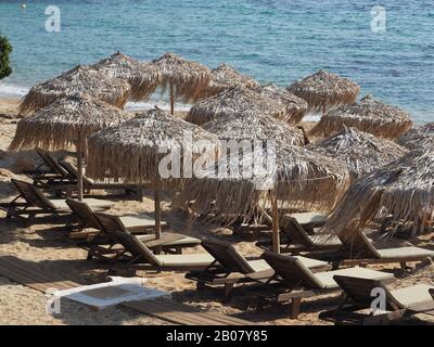Menschenleere Sonnenliegen aus Holz mit Strohschirmen an einem Strand in Evia, Griechenland. Blaues Meer dahinter. Stockfoto