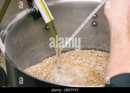 Ein Mann gießt Wasser in einen Tank, in dem Bodenmalz liegt. Messung der Wassertemperatur mit einem Thermometer. Der Prozess der Herstellung von hausgemachtem Bier Stockfoto