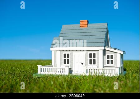 Miniaturhaus mit weißem Pickelzaun, der in einem grünen Rasen vor einem blauen Himmelshorizont steht Stockfoto