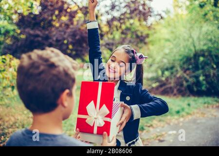 Fröhlicher kleiner Kinderjunge schenkt lächelnden Mädchen eine Geschenkbox, was eine Überraschung macht. Fröhliches kleines Mädchen, das ein Geschenk in einem roten Kasten öffnet. Stockfoto