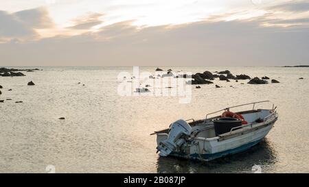 Emotionaler Meereseinbruch im Morgengrauen an einem kleinen Strand mit bewölktem Himmel und einem am Ufer verankerten Fischerboot mit einem Seil, das sich in flachem Wasser bewegt Stockfoto