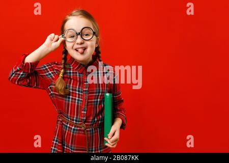 Lustige fröhliche Kinder in Brille und mit einem Buch auf rotem Hintergrund. Kleines Mädchen, das sich herumtäuscht und Zunge zeigt. Stockfoto