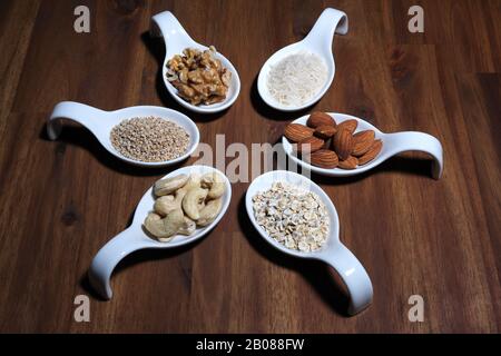 Zutaten für Vegan-Milch zur Selbstherstellung, auf einer Holzplatte in weißen Schüsseln, Mandel, Nussbaum, Reis, Haferflocken, Cashewnüsse, Sesams angeordnet Stockfoto