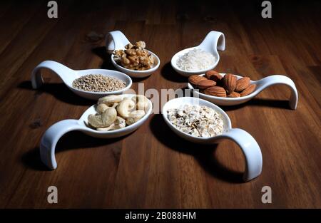 Zutaten für Vegan-Milch zur Selbstherstellung, auf einer Holzplatte in weißen Schüsseln, Mandel, Nussbaum, Reis, Haferflocken, Cashewnüsse, Sesams angeordnet Stockfoto